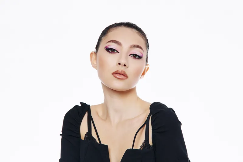 Beauty model with creative makeup in soft pink and bronze hues poses confidently in black attire. Studio portrait on white background for fashion and beauty editorial.