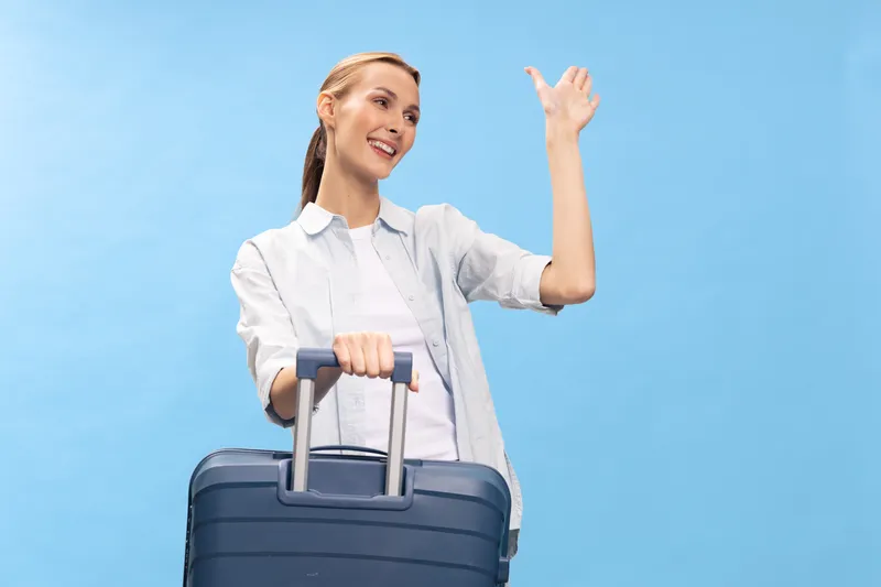 Young woman with suitcase waves hand and smiles while standing against blue background. Concept of travel, vacation, tourism, and holiday preparation with casual outfit and positive mood.