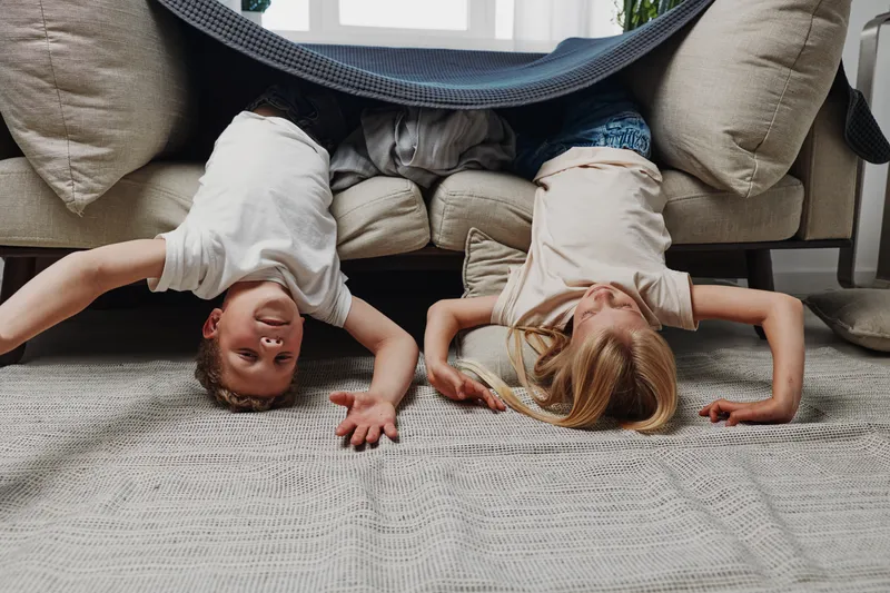 Two children lie upside down on the floor under a blanket draped over a couch. The playful scene shows fun and imagination in a cozy living room environment.