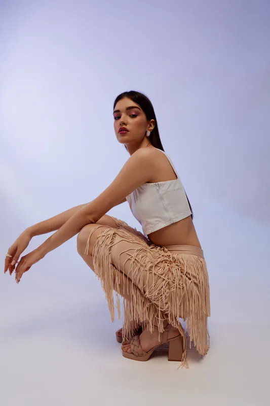 Young woman in a white crop top and beige fringed pants poses in a studio with a neutral background. Concept for woman beauty fashion and modern style.