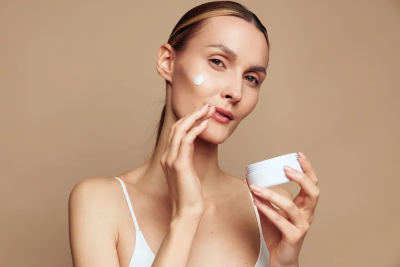 Young woman applying white face cream on cheek with fingers. Close-up portrait on beige background. Concept of skincare, beauty routine, and self-care with natural lighting.
