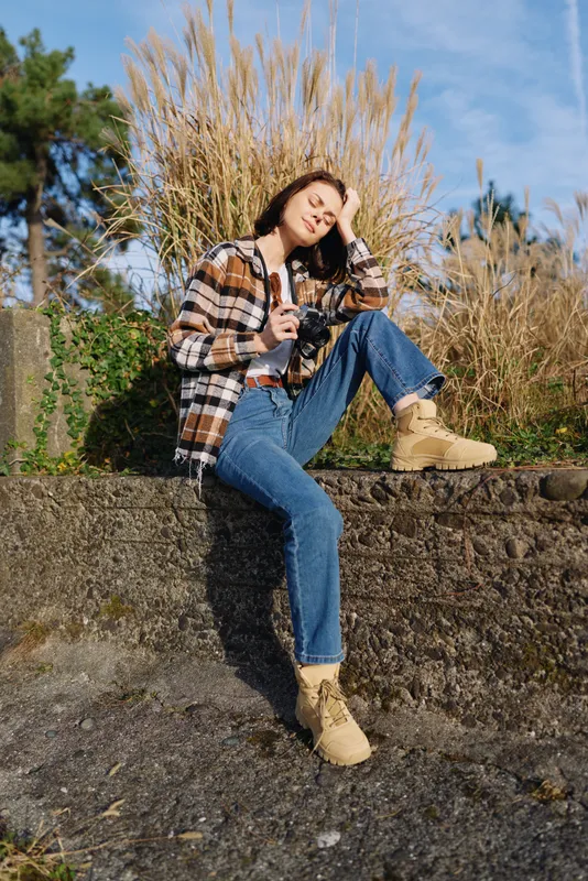 Young woman sits on a stone wall outdoors, wearing a plaid shirt, jeans and boots, enjoying sunlight and fresh air in a casual fashion moment.