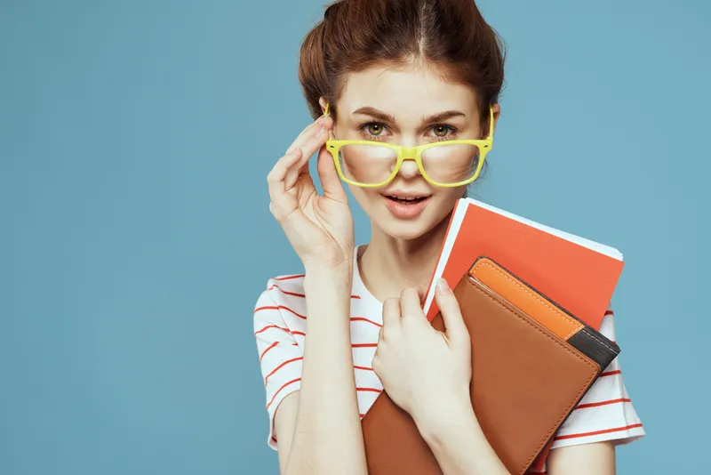 Студентка с книгами в руках на синем фоне, в жёлтых очках и с модельной причёской, кадрированный вид