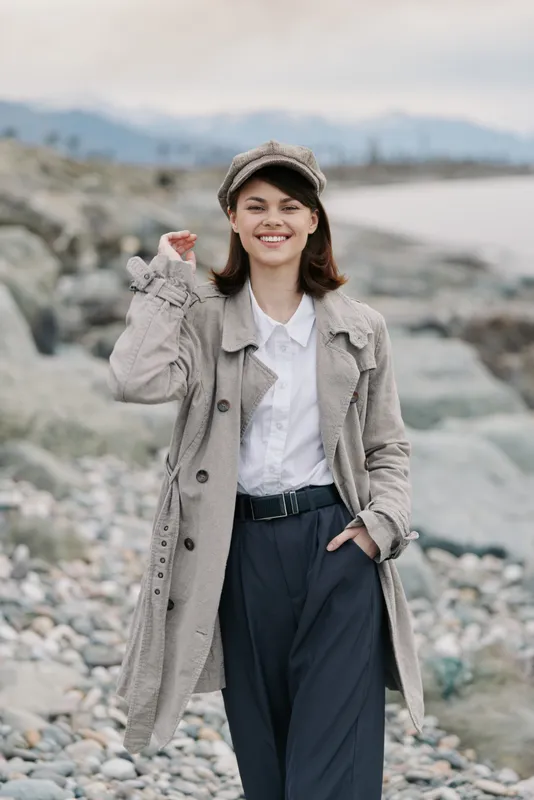 Smiling young woman in textured coat and cap stands on rocky beach with mountains and sea in background. Casual fashion style and natural outdoor setting create relaxed mood.