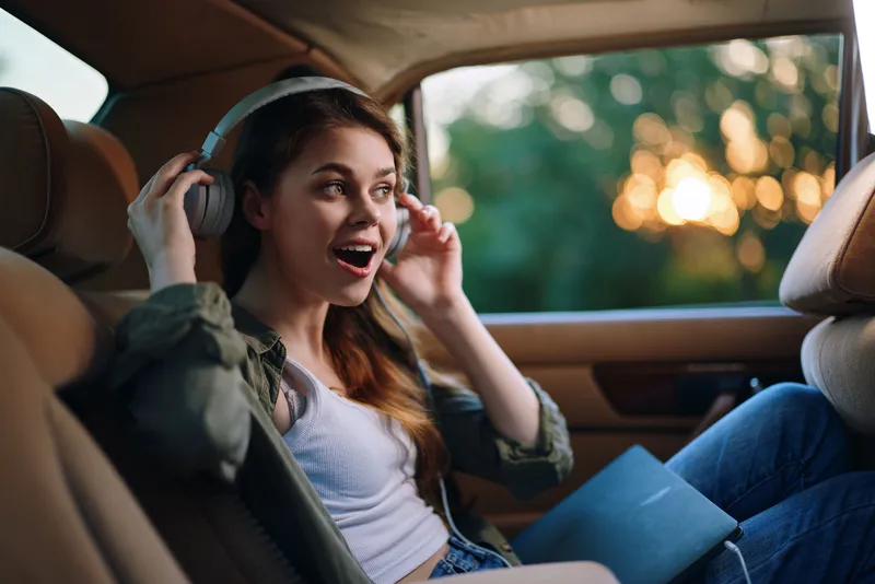 Young woman enjoying music in a car, smiling with headphones on, casual attire, sunlight filtering through the window, conveying joy and freedom.