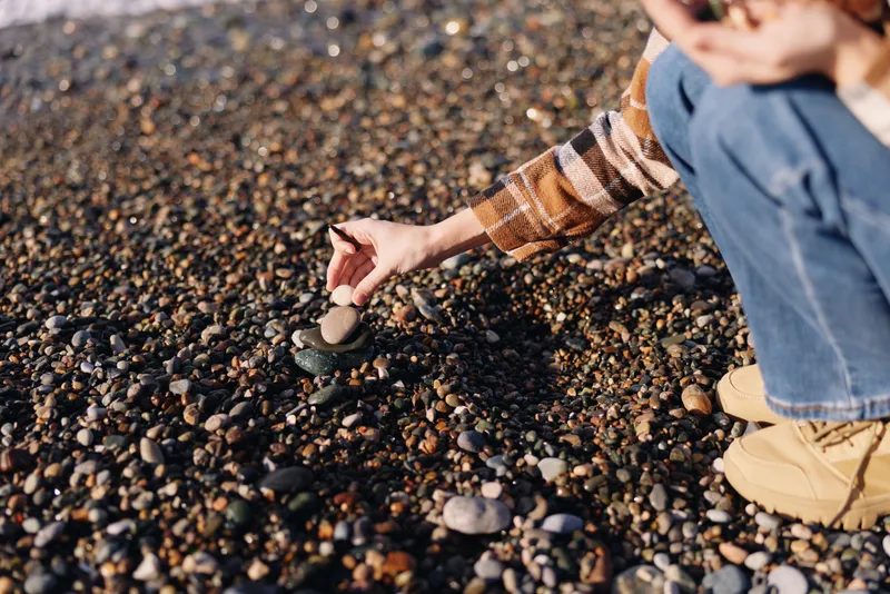 Pebble beach, rock in hand, outdoor exploration, casual attire, kneeling on pebbles to collect a stone during a calm coastal moment, highlighting texture and coastal scenery