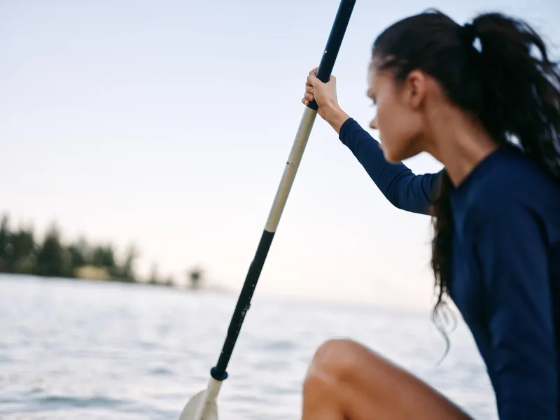 A focused rower grips a paddle while gliding across calm water, emphasizing strength, balance, and motion in a tranquil outdoor scene under soft light.