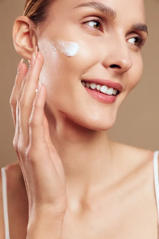 Young woman applies moisturizing cream on clean glowing skin with fingers, shows skincare routine, smiles with natural makeup, isolated on beige background closeup.
