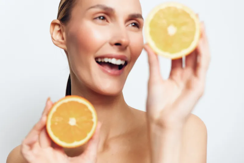 woman, smiling, orange, citrus, skincare, beauty, wellness, natural portrait, young woman holds orange slices by face, soft light, fresh glow, clean white background