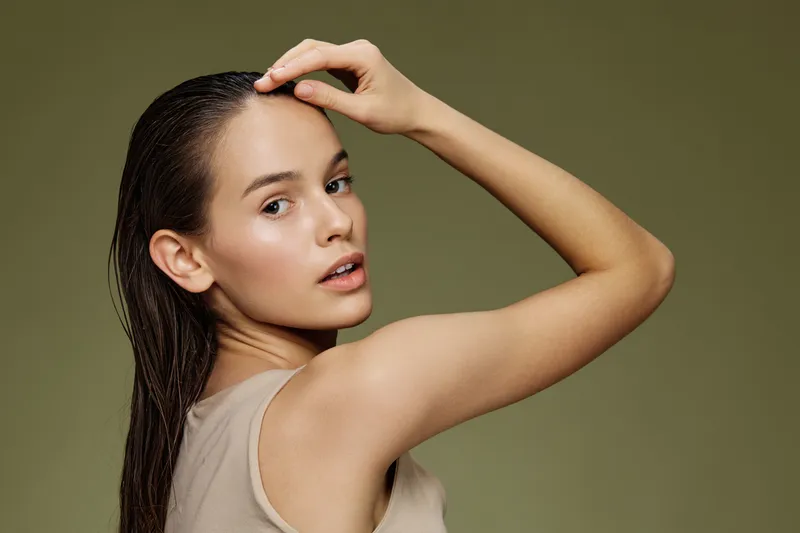 Young model with long hair, wearing a neutral colored outfit, showcasing a serene expression against a soft green background, emphasizing natural beauty and skincare concepts