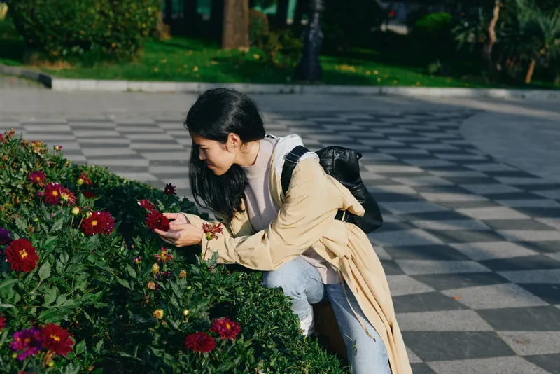 Woman crouching beside vibrant flowers in a park, inspecting blossoms with a backpack on her shoulder, casual coat, serene outdoor scene showcasing nature, leisure activity and spring mood