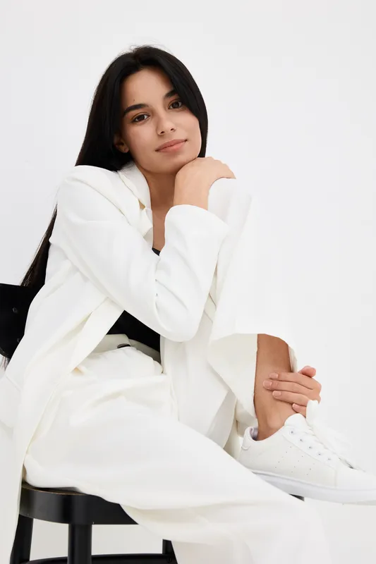Young woman in white suit sitting thoughtfully on black chair, with long dark hair, casual sneakers, minimalist studio background, natural expression.