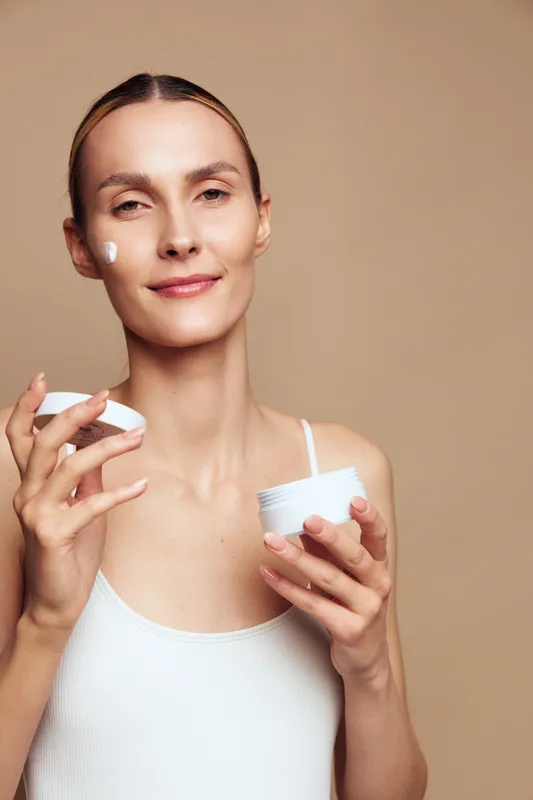 A young woman with clear skin applies white cream on her cheek while holding an open jar in the other hand. She wears a white tank top against a beige background. Concept for skincare and beauty.