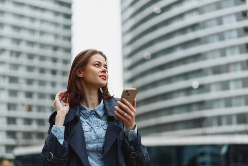 Young woman in an urban setting checks her smartphone, wearing a denim shirt and navy coat, looking up with a confident expression amid tall modern buildings.