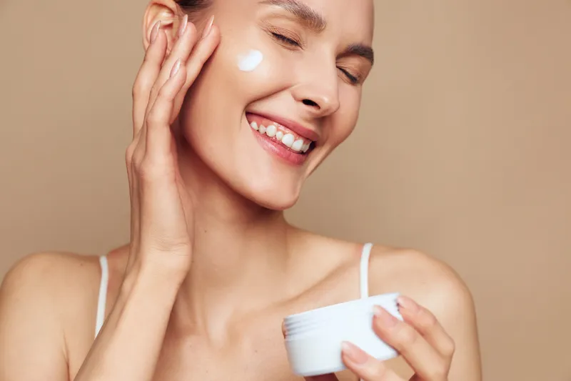 Smiling woman applying white face cream on cheek, holding jar in hand. Close-up portrait with closed eyes on beige background. Concept of skincare, beauty, and self-care routine.