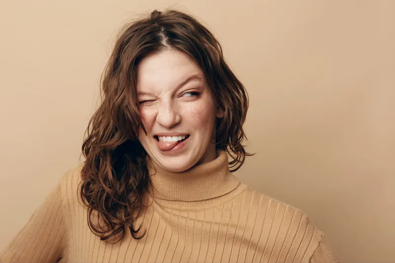 Young woman with curly hair wearing beige sweater makes funny face with tongue out and wink. Studio portrait on neutral background. Concept of humor, playfulness, and casual style.