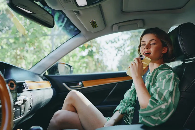 A cheerful woman sits in the car's driver seat, enjoying a lollipop, relaxed and ready for a carefree journey.