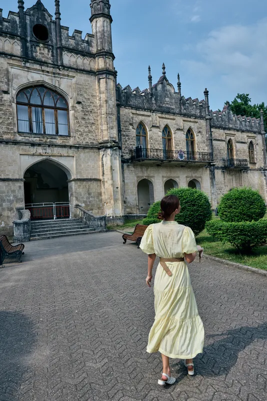 Castle grounds with a graceful woman in a pale dress walking along a stone pathway toward a grand estate, featuring gothic architecture and manicured gardens.