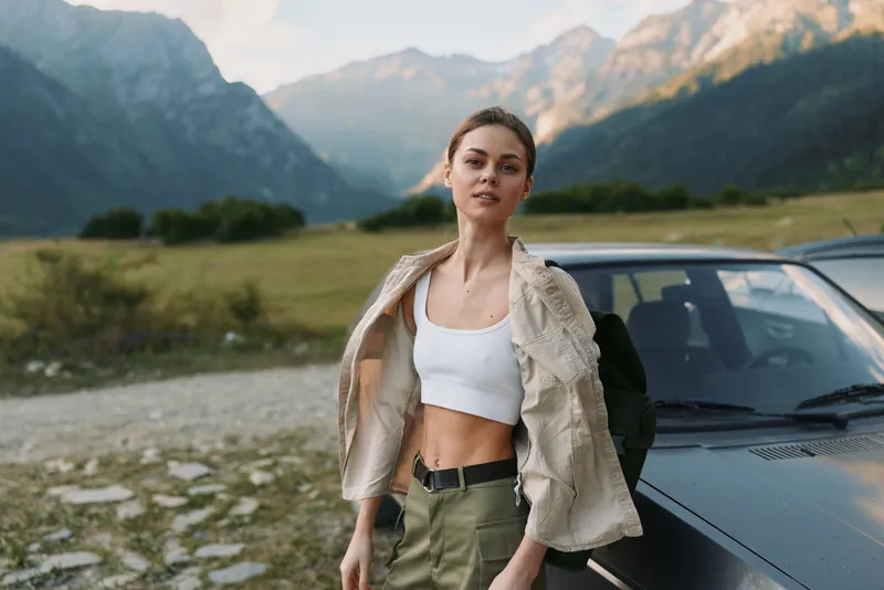 Young woman stands beside a car in a rugged mountain landscape, unbuttoning a light jacket and enjoying a relaxed, adventurous moment.