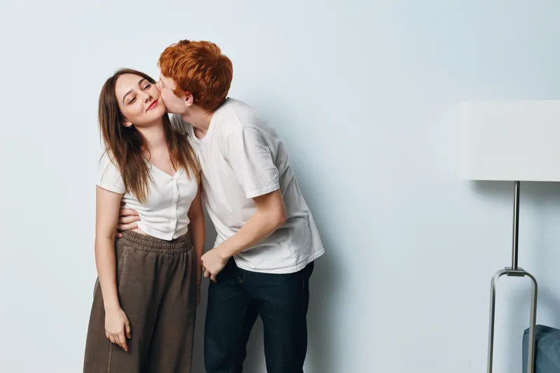 Young couple, man kissing woman on cheek for love, affection and romance. Casual clothing, happy relationship with intimacy and tender moment at home interior with lamp.
