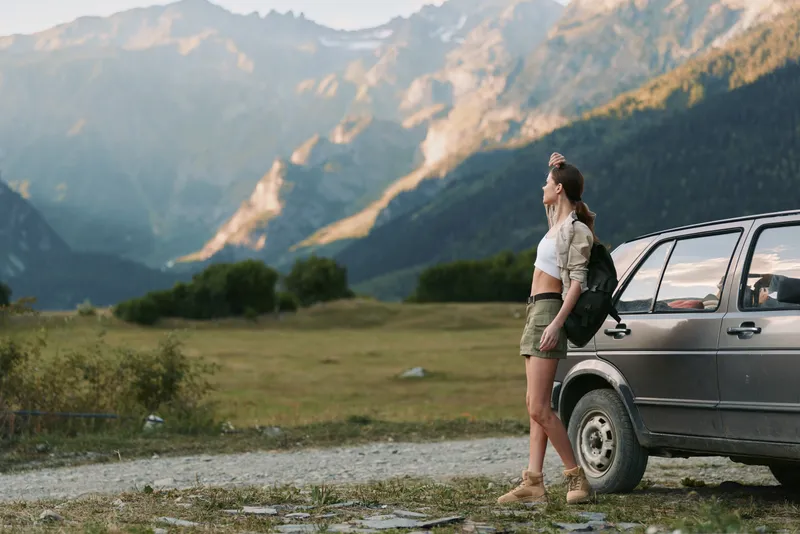 Young woman with backpack stands beside a car in a mountain valley, taking a moment to soak in the rugged scenery during a daytime road trip