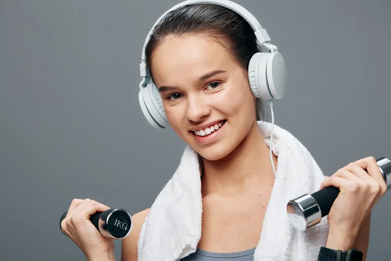 Woman fitness and sport in a gym setting, lifting dumbbells with a towel over her shoulders, wearing headphones, showing strength, focus, and motivation for workouts.