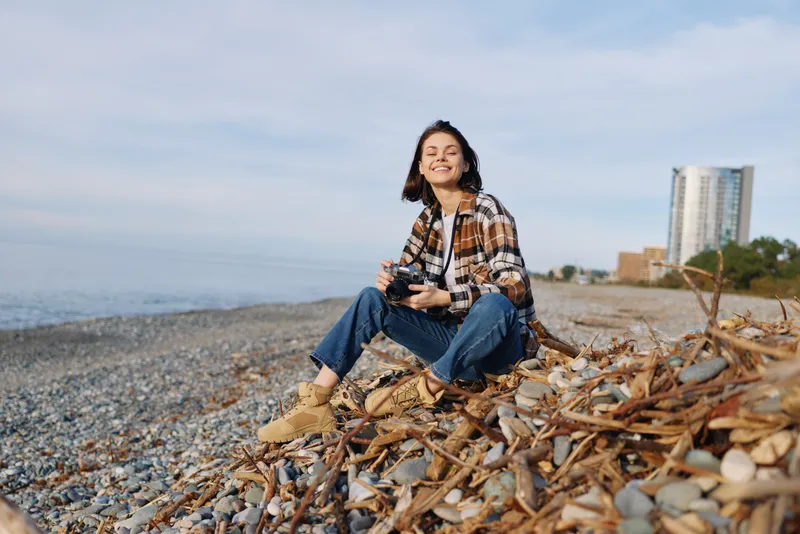 Woman on a pebble beach with a camera, sitting and smiling in casual attire, relaxed mood by the sea with distant buildings and clear sky.