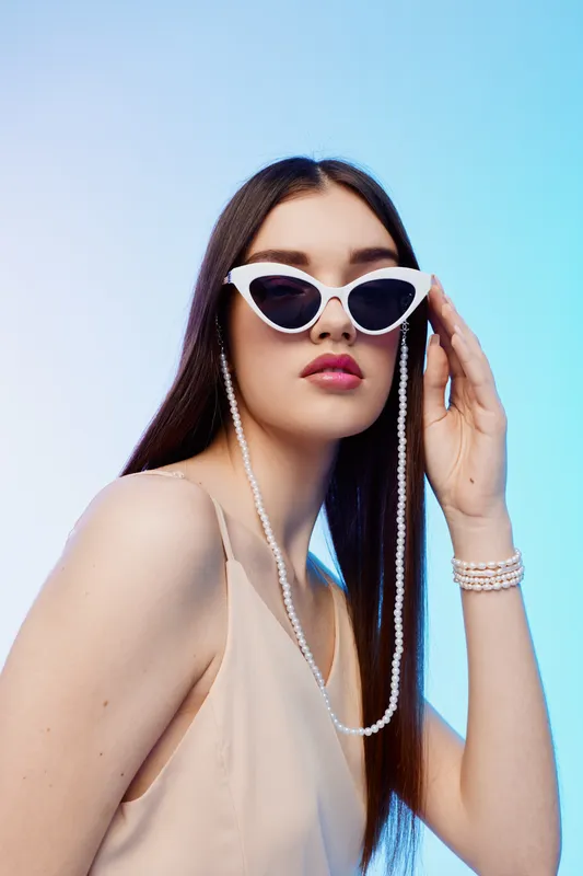 Young woman in white sunglasses and beige top poses against light blue background. Long hair and pearl accessories highlight beauty and fashion concept.