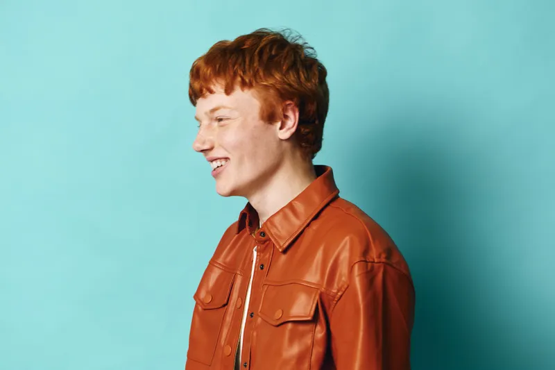 Young man with red hair wearing brown leather jacket smiling and looking sideways with bright blue background, casual portrait with happy expression and trendy style.