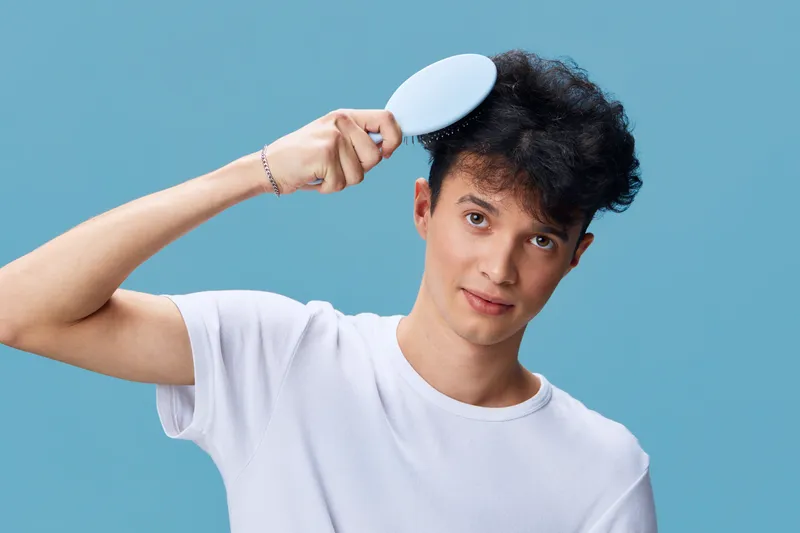 Young man brushing curly hair against a blue background wearing a casual white t-shirt, showing natural grooming and personal care with a focused expression.