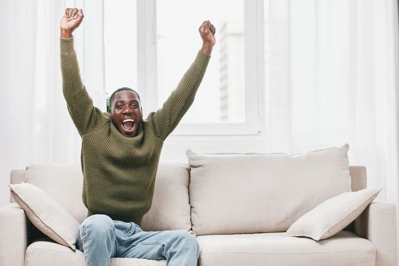 Happy Black man celebrating success in a cozy living room, wearing a green sweater, embodying joy and excitement with a bright atmosphere