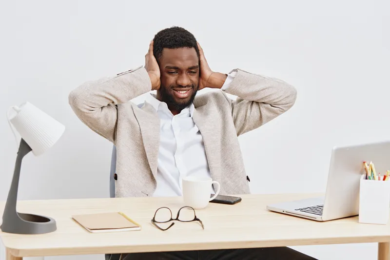 Smiling man in beige jacket and white shirt sitting at desk with laptop, mug, glasses, and notebook in bright modern office environment. People lifestyle concept