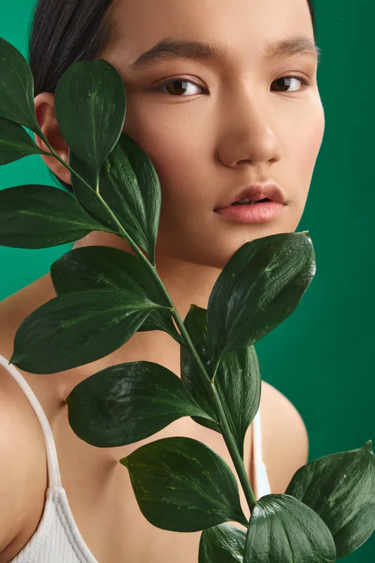 asian woman poses with lush green leaves in a studio setting, close up portrait, serene expression, soft lighting, vibrant background conveys natural and fashionable mood.