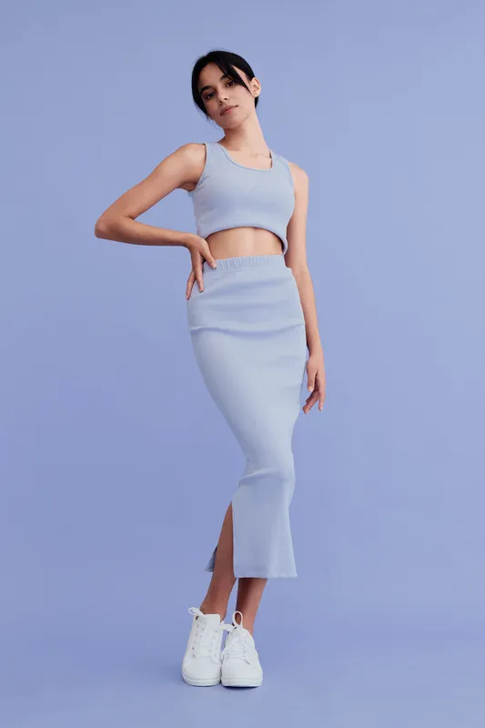 young woman in light blue sleeveless crop top and matching skirt stands confidently against pastel purple background, wearing white sneakers, casual modern outfit style