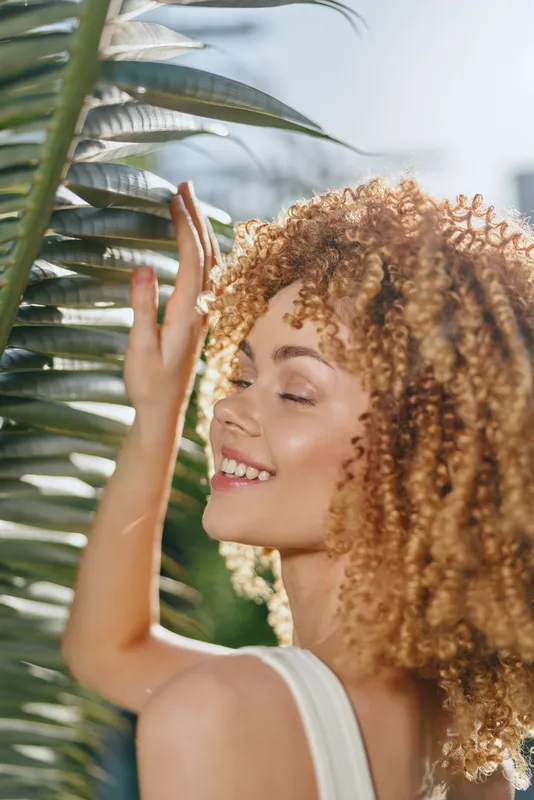 Smiling woman with curly hair enjoying sunlight near green palm leaves, radiant skin, peaceful expression, natural beauty captured outdoors.