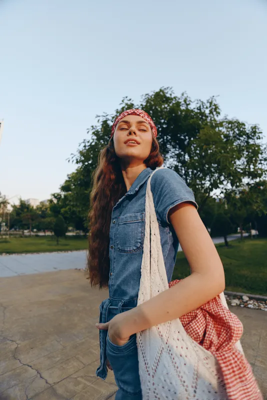 Young woman in denim jumpsuit with red bandana carrying crochet bag outdoors on sunny day with green trees and clear sky background