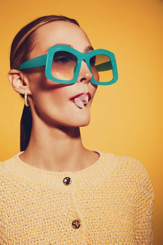 Stylish woman in oversized turquoise sunglasses and yellow sweater poses against orange background. Fashion portrait with bold accessories and modern casual outfit concept.