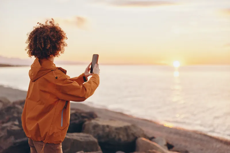 Woman lifestyle portrait at sunset by ocean, standing on seashore with smartphone, curly hair visible, casual jacket, warm golden light, peaceful travel outdoors on summer evening.