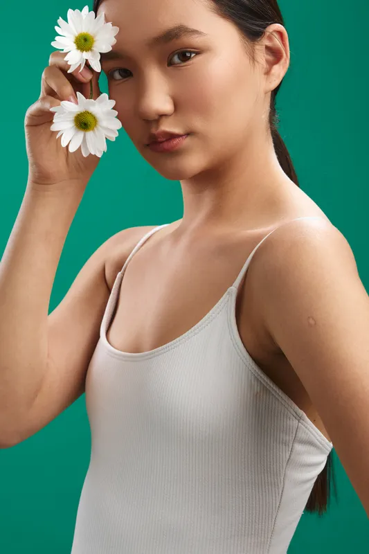 asian woman wearing a light tank top stands before a vibrant green backdrop, softly touching white daisies near her face, conveying fresh beauty, calm confidence, and natural elegance.