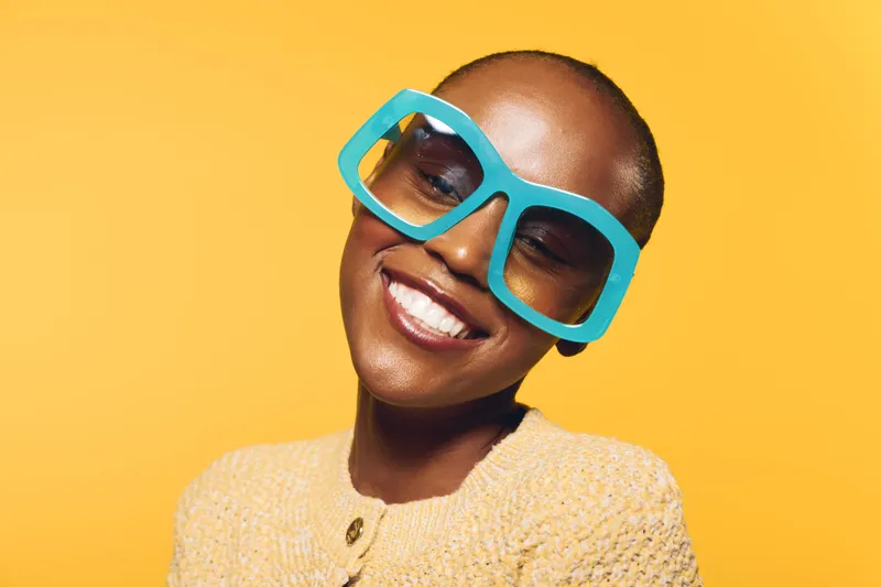 Smiling dark skinned African woman wearing oversized blue sunglasses and yellow sweater poses against yellow background. Portrait of happy black female with short hair and bright mood.