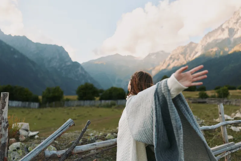 Cheerful woman outdoors, arms outstretched, blanket draped over shoulders, sunlit valley with towering mountains, relaxed lifestyle moment, fresh air, natural colors, serene backdrop, hopeful mood