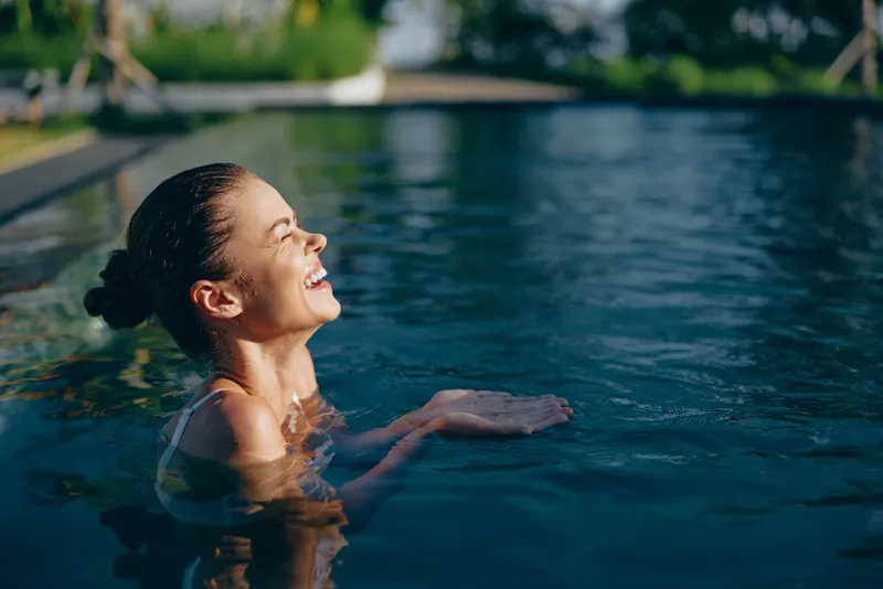 A joyful woman rises from a pool, laughing with head tilted back. Clear water frames her relaxed pose, suggesting carefree summer leisure and personal wellness in a sunny outdoor setting.