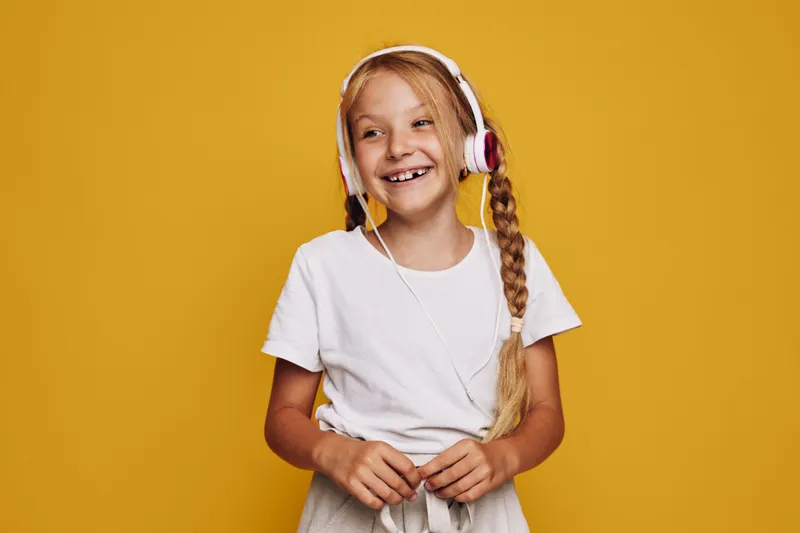 Smiling blonde girl with long braid wearing white t-shirt and headphones enjoying music against yellow background. She looks happy and relaxed with a joyful expression.