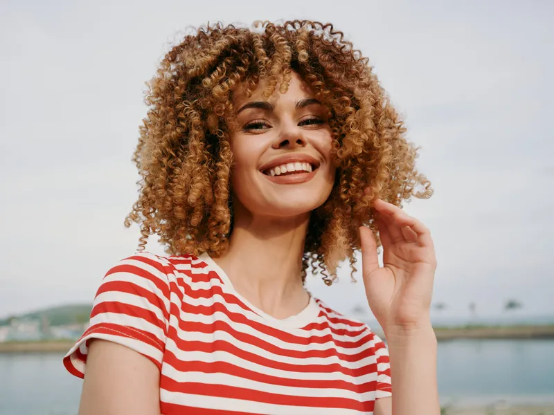 Portrait of a joyful young woman with curly hair wearing a red and white striped shirt, standing outdoors by a calm body of water under soft daylight.