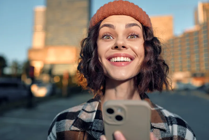 Woman smile phone urban beanie, candid portrait of a joyful young adult holding smartphone in a modern city at golden hour glow, authenticity and mindful living with emotional storytelling vibe.