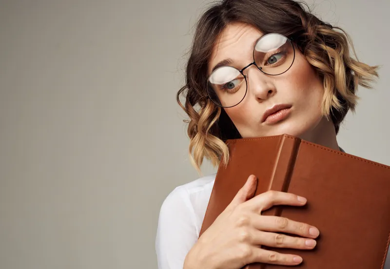 Романтичная женщина в очках на сером фоне с коричневой книгой в руках