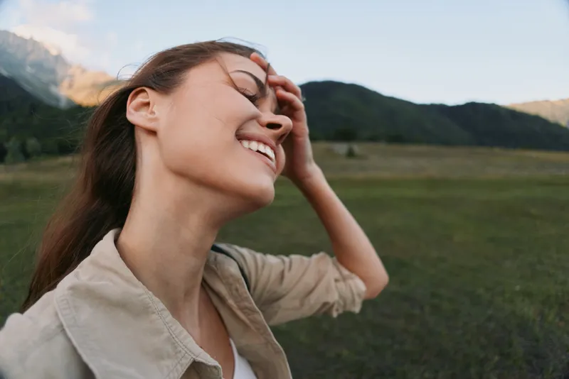 Smiling woman outdoors in a grassy field, wind in hair, basking in sunlight. Natural lifestyle portrait conveying wellness, freedom, and relaxed joy.