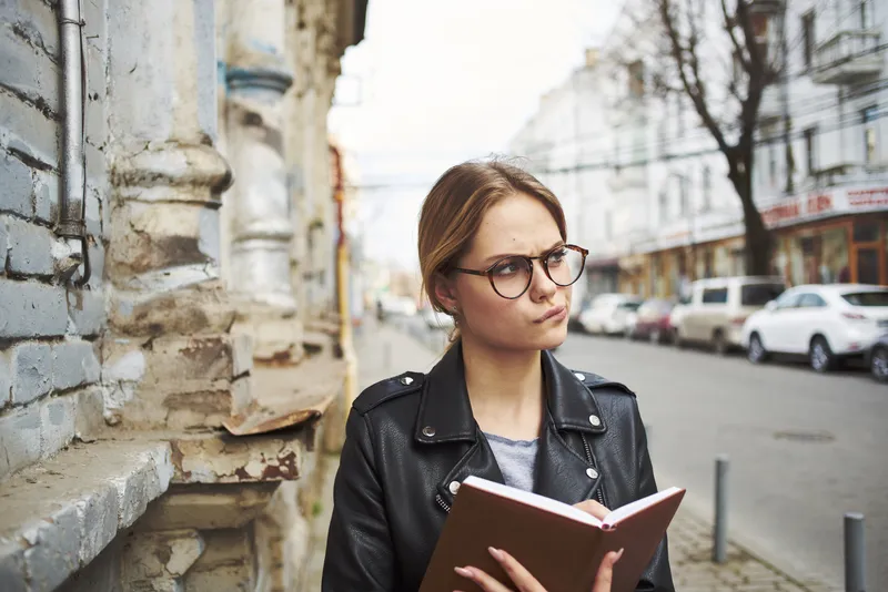 Счастливая женщина в кожаной куртке с книгой в руке идет по улице в городе