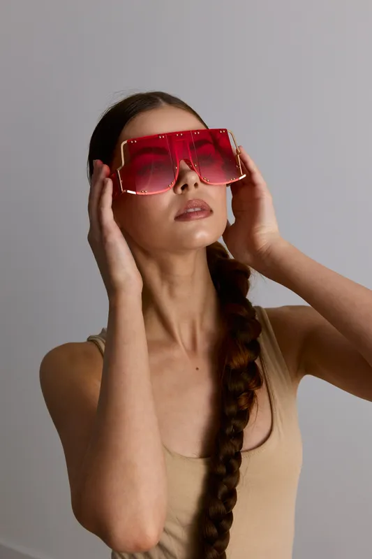 Woman wearing oversized red sunglasses with braided long hair and neutral beige top against gray background, showcasing modern fashion and bold summer style.