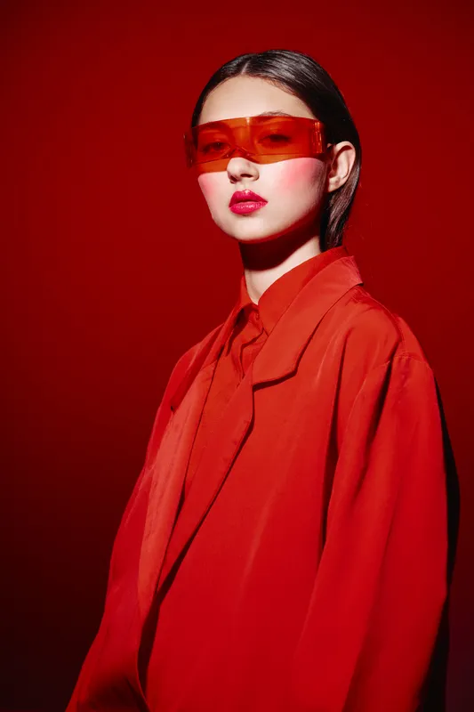 Fashion model wearing a vibrant red oversized blazer and tinted orange sunglasses poses confidently against a bold red background for a creative beauty and fashion concept shot.